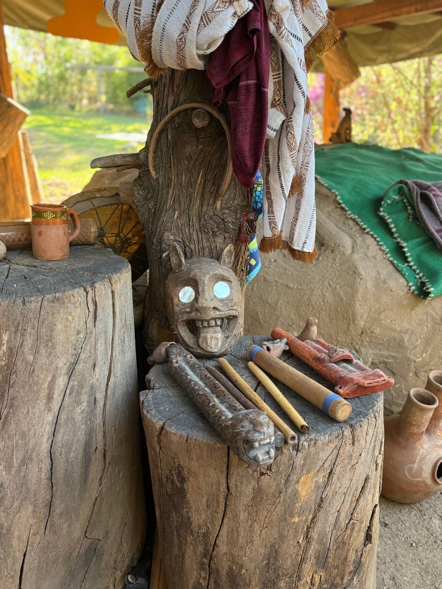 Carved mask, clay flutes, and woven textiles laid out on tree-stump tables for the ceremony